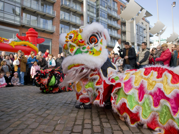 Ein lebhaftes chinesisches Neujahrsfest in Amsterdam mit einer Löwen-Tanzvorstellung vor einer Zuschauermenge, darunter einige, die das Ereignis fotografieren, vor einer Kulisse aus Gebäuden, Laternenmasten und einem klaren blauen Himmel.