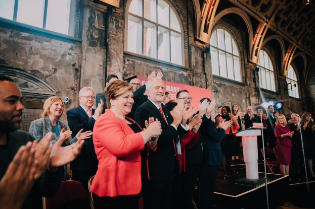 Eine Gruppe von Menschen steht vor einer Menge, klatscht in die Hände und feiert, mit einem Podium mit Mikrofon auf der rechten Seite und einem Banner im Hintergrund, das 'Covid-19 New York City Mayor Bill de Blasio' lautet.
