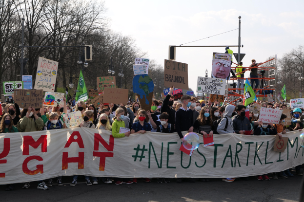 Eine große Gruppe von Menschen marschiert auf einer Straße, hält ein Transparent mit der Aufschrift 'Menschliche Rechte' und trägt Masken oder trägt Schilder; Bäume, Laternenpfähle und ein klarer blauer Himmel sind im Hintergrund zu sehen.