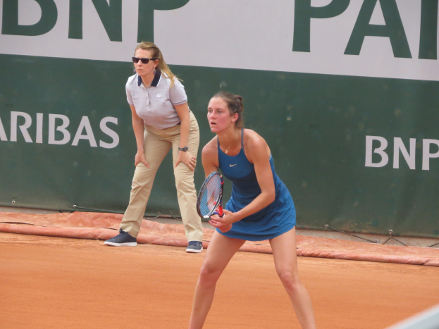 Zwei Frauen beim Tennis auf einem Sandplatz, wobei eine in einem blauen Kleid mit einem Schläger in der Hand im Vordergrund steht und eine andere dahinter steht, während ein grüner Banner mit Text im Hintergrund zu sehen ist.