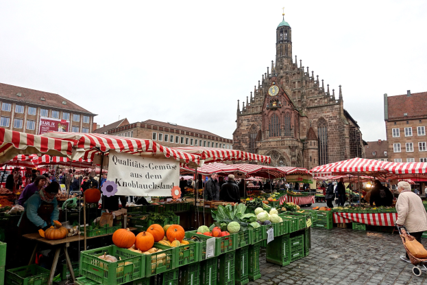 Ein belebter Markt in Nürnberg, Deutschland, der verschiedene Früchte und Gemüse zeigt, mit Menschen und Zelten, vor Gebäuden und einem Uhrturm unter einem sichtbaren Himmel.