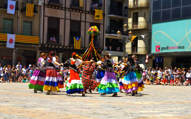 Eine Gruppe von Frauen in bunten Kleidern tanzt auf der Straße, umgeben von einer Menschenmenge, mit Gebäuden, Geländern, Balkonen, Fahnen, einem Laternenpfahl und einer Tafel mit Aufschrift im Hintergrund.