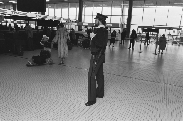 Polizeibeamter in Uniform mit Mütze, der eine Waffe hält und in einem Flughafen-Terminal steht, umgeben von Menschen und Gepäck, mit Gepäckwagen, Säulen, Informationsanzeigen und Glastüren im Hintergrund.