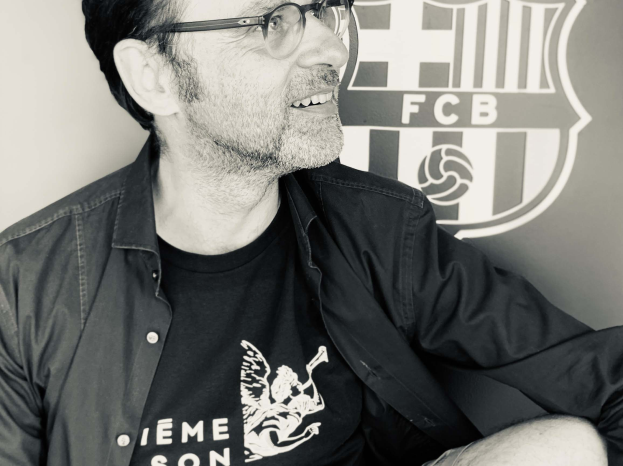 Ein Mann in einer Brille und einem schwarzen Hemd lächelt vor einem schwarz-weißen FC Barcelona-Logo an einer Wand.