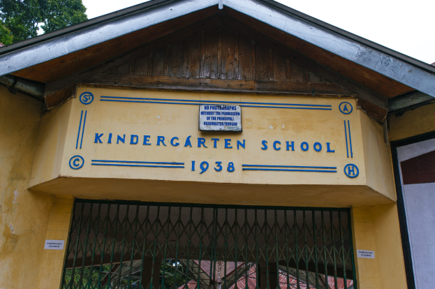 Gelbes Gebäude mit einer "Kindergarten-Schule"-Schilder und einer Wandtafel, einem Gitter unten und Bäumen im Hintergrund vor einem klaren blauen Himmel.