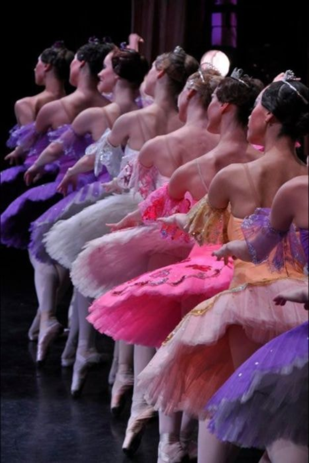 Eine Gruppe von Balletttänzerinnen in rosafarbenen Tutus sitzt auf einer Bühne, beleuchtet von einem Licht im Hintergrund, und führt synchronisierte Bewegungen aus.