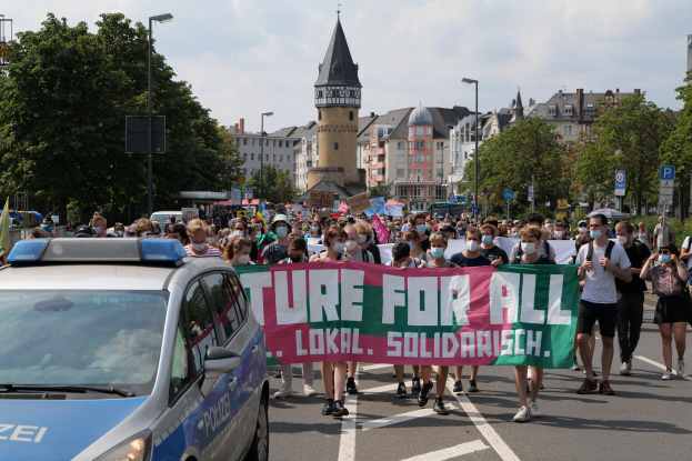 Eine große Gruppe von Menschen marschiert mit einem Banner, auf dem "Lokale Solidarität" steht, eine Straße entlang, neben einem Polizeiwagen geparkt, Bäume und Gebäude im Hintergrund unter einem bewölkten Himmel.