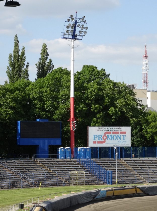 Rennstrecke beleuchtet von Oberlichtern, umgeben von Gras, Geländern, Stufen, einem Lichtmast, einer Tafel, Bäumen, Gebäuden, Türmen und einem bewölkten Himmel.