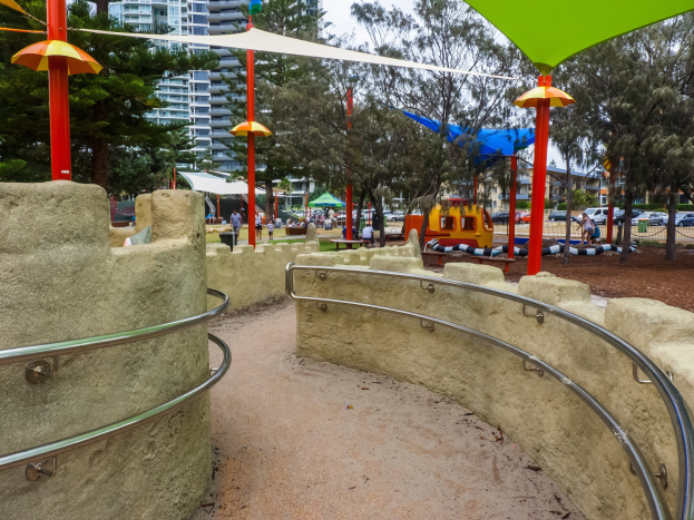 Spielplatz mit einem grünen und gelben Schirm, umgeben von Zäunen, Pfählen, Zelten, Bäumen, Fahrzeugen, Menschen und Gebäuden unter einem klaren blauen Himmel.