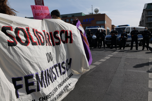 Gruppe von Menschen marschiert auf einer Straße, die eine 'Solidarität und Feminismus'-Fahne hält, mit parkenden Autos, Gebäuden, einer Schüsselantenne und einem klaren blauen Himmel im Hintergrund.