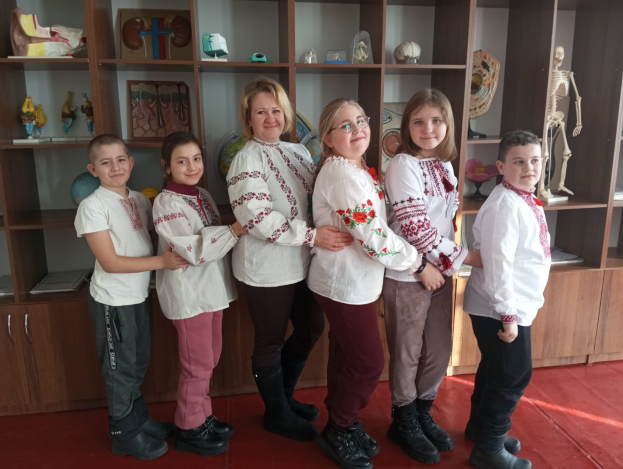 Gruppe von Kindern in traditioneller ukrainischer Kleidung vor einem Bücherregal mit Spielzeug, einem Skelett und anderen Gegenständen.