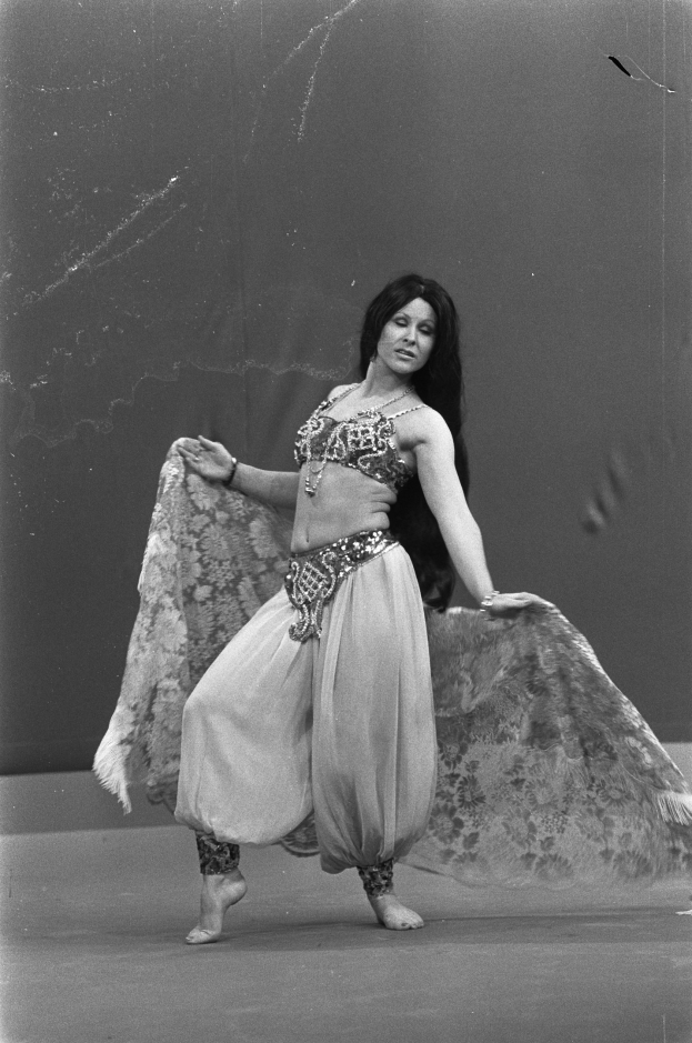 Eine Frau in einem langen, gemusterten Bauchtanzkleid tanzt mit geschlossenen Augen und ausgestreckten Armen vor einer Wand