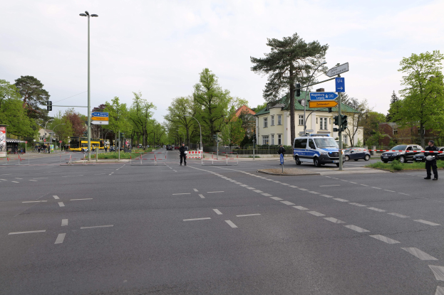 Rundverkehr mit einem auf der Seite parkenden Polizeiwagen, fahrende Fahrzeuge, Fußgänger auf dem Gehweg, Verkehrsampeln, Bäume, Gebäude mit Fenstern und bewölkter Himmel.