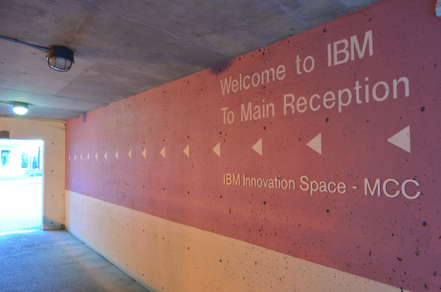 Eine Wand mit dem Text "Willkommen bei IBM" beleuchtet von Deckenlampen, mit einer Tür im Hintergrund beschriftet mit "IBM Innovation Space MCC."