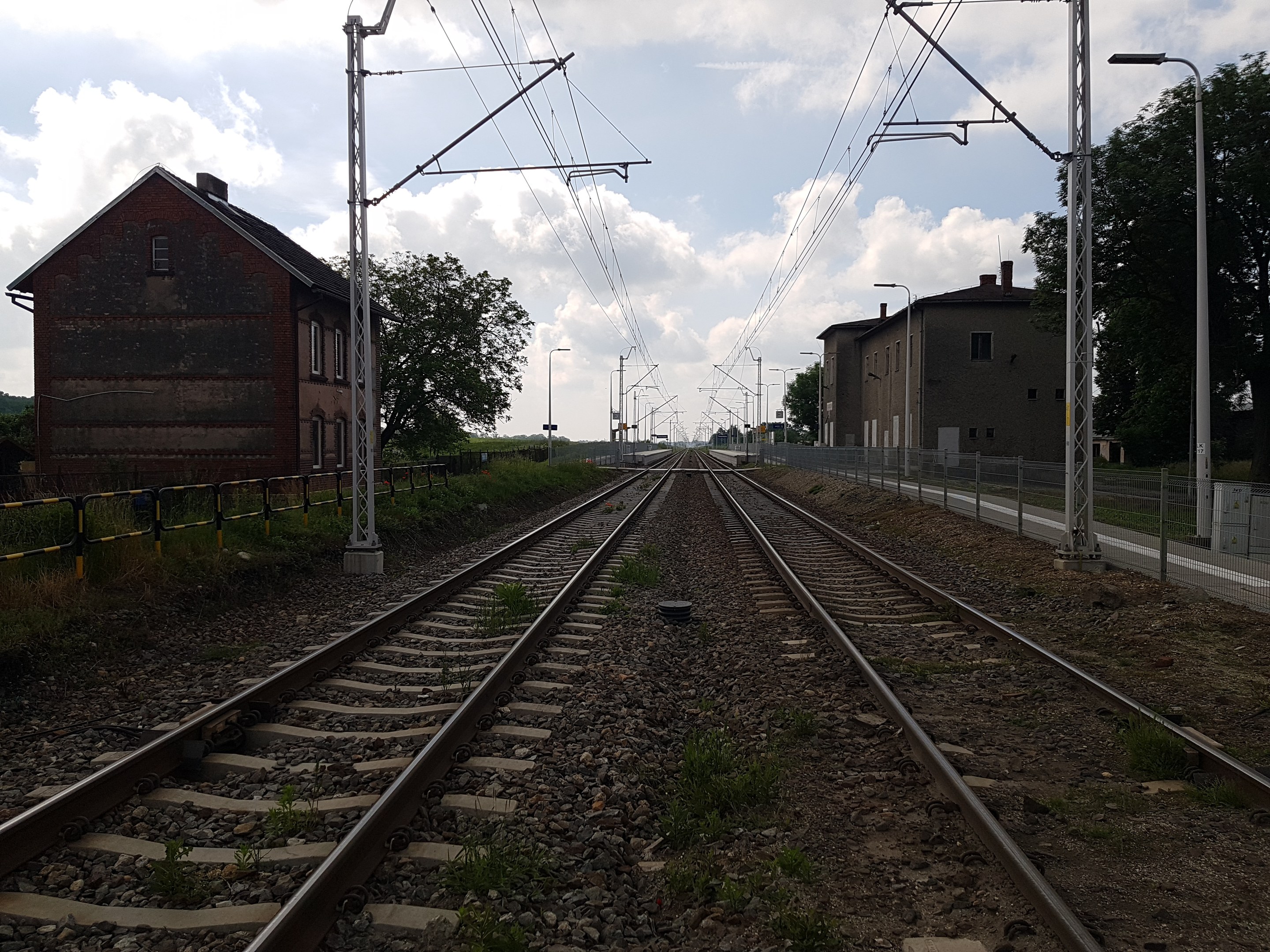 Eine Schiene mit Oberleitungen, benachbarte Gebäude, Straßeninfrastruktur, Vegetation und ein bewölkter Himmel.
