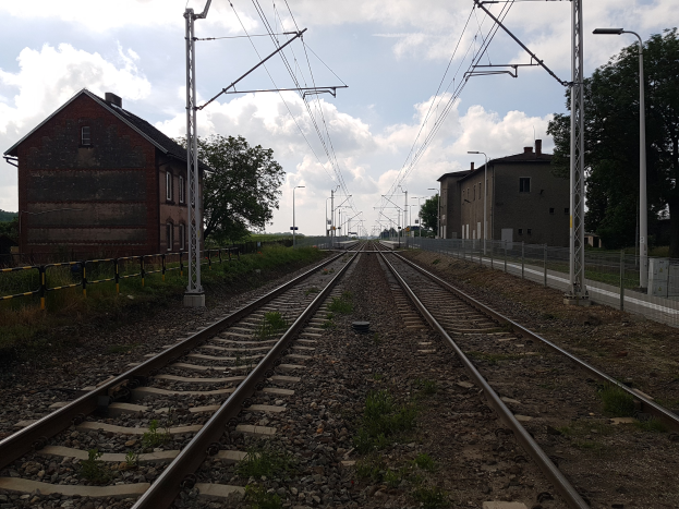 Eine Schiene mit Oberleitungen, benachbarte Gebäude, Straßeninfrastruktur, Vegetation und ein bewölkter Himmel.