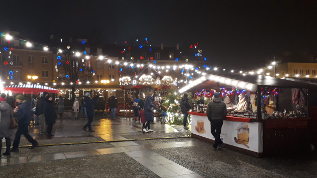 Ein lebendiger Weihnachtsmarkt am Abend in einer Stadt, mit Menschen, die herumlaufen, Ständen, die mit Lichtern und Dekorationen geschmückt sind, Gebäuden mit Fenstern, Bäumen und einem Himmel, der mit festlichen Lichtern erleuchtet ist.