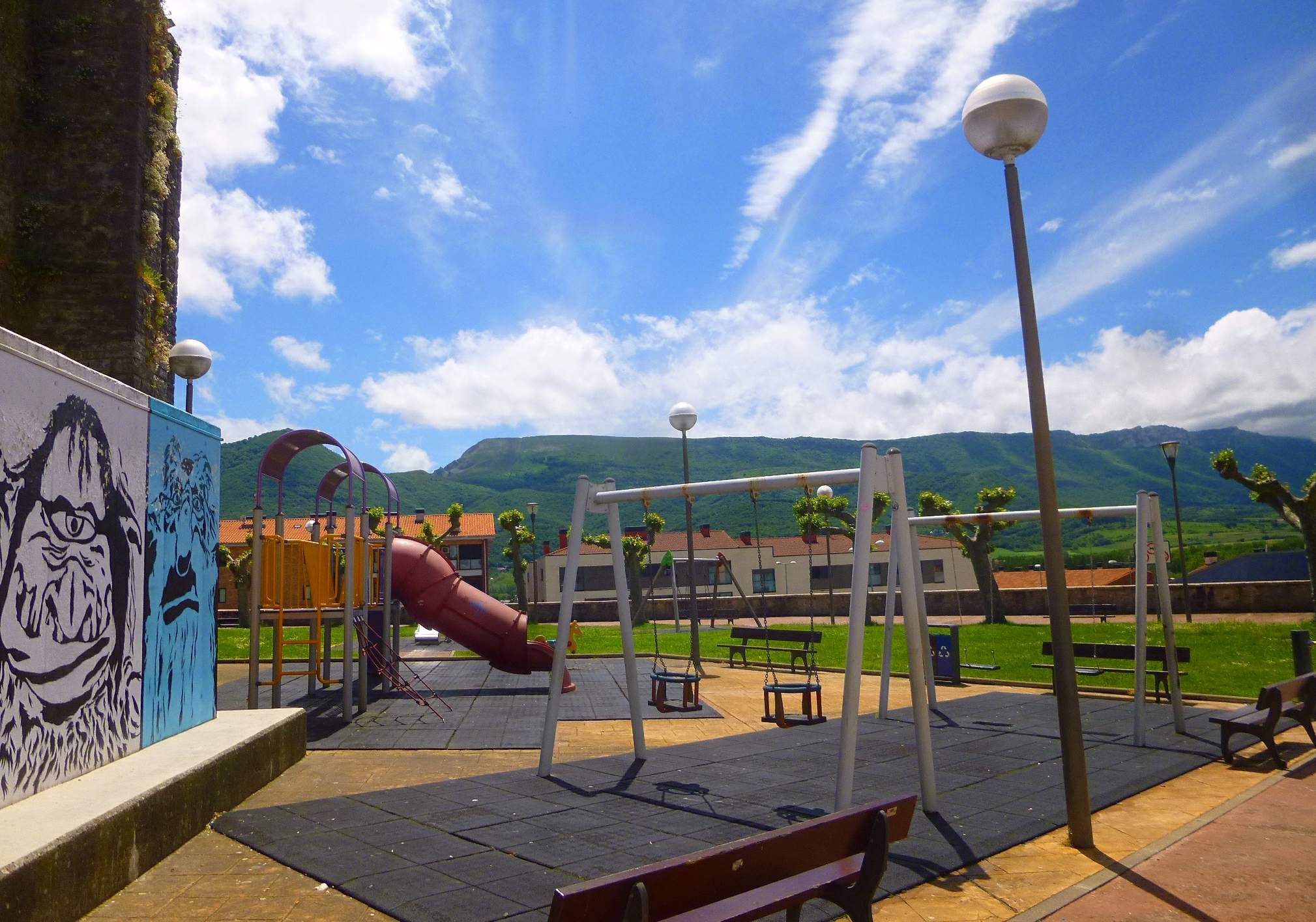 Ein Spielplatz mit Schaukeln, einer Rutsche, Bänken, Pfosten, Laternen, einer bemalten Wand, Gras, Bäumen, Gebäuden, Hügeln und einem bewölkten Himmel im Hintergrund.