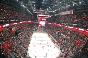 Eine große, hell erleuchtete Arena voller jubelnder Zuschauer, die ein Hockey-Spiel sehen, mit einem zentralen Bildschirm über der Eisfläche.