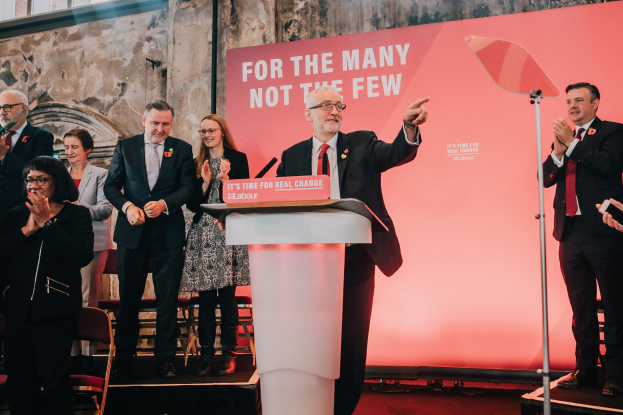 Ein Mann in einem schwarzen Blazer, weißem Hemd und roter Krawatte spricht an einem Podium mit einem Mikrofon vor einem klatschenden Publikum, mit einem Banner an der Wand dahinter und einer Lampe rechts daneben.