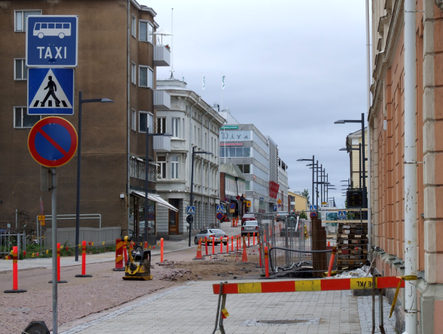 Stadtstraße mit Gebäuden, Straßenlaternen, Schildern, Verkehrsleitkegeln, Fahrzeugen, Absperrpoller, Bäumen und einer Baustelle mit Verkehrszeichen unter einem bewölkten Himmel.