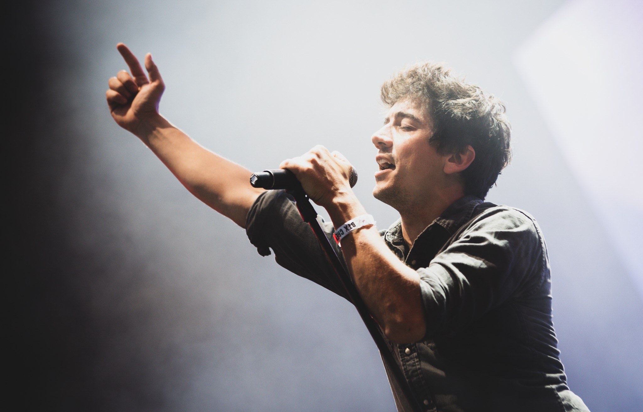 Ein Mann in schwarzem Hemd und Jeans singt leidenschaftlich in ein Mikrofon auf einer hell erleuchteten Bühne, seine Augen geschlossen und das Haar unordentlich gestylt, während das Publikum im Hintergrund applaudiert.