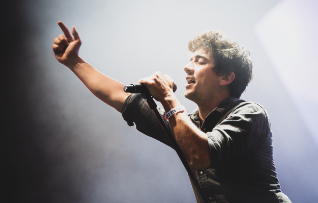 Ein Mann in schwarzem Hemd und Jeans singt leidenschaftlich in ein Mikrofon auf einer hell erleuchteten Bühne, seine Augen geschlossen und das Haar unordentlich gestylt, während das Publikum im Hintergrund applaudiert.