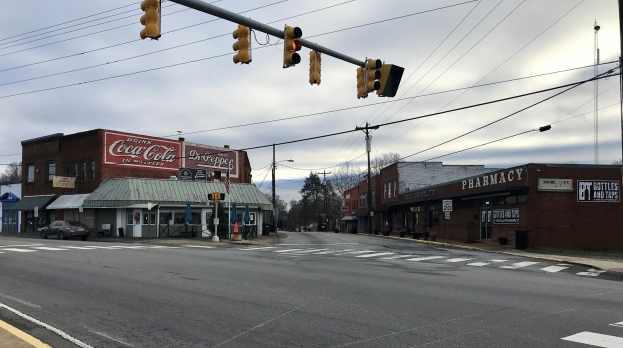 Stadtstraßenkreuzung mit Ampel, Fahrzeugen, Gebäuden mit Schildern, Strommasten mit Drähten, Bäumen und einem bewölkten Himmel.