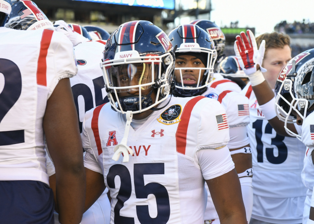 Gruppe von Männern in Football-Uniformen auf einem Feld mit Helmen und Handschuhen, mit einer unscharfen Menge, einer Anzeigetafel mit "Auburn vs Virginia Tech - 11/28/15 College Football Pick, Odds, and Prediction" und Himmel im Hintergrund.