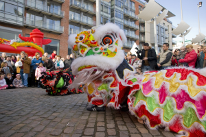 Ein lebendiges chinesisches Neujahrsfest in Amsterdam mit einer Löwendance-Vorführung und einer Zuschauermenge, einige halten Kameras, vor einem Hintergrund aus Gebäuden, Laternenmasten und einem klaren blauen Himmel.