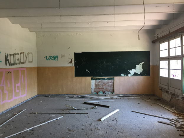 Leerer Klassenraum mit graffiti-bedeckten Wänden, einer zentralen Tafel, Glasfenstern auf der rechten Seite, einem mit Schutt übersäten Boden und einer sichtbaren Decke.