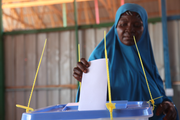 Frau in blauem Kopftuch, die ein Blatt Papier hält und vor einer Kiste mit herausragenden Stöcken steht, vor einer Wand und einem Schuppen im Hintergrund.