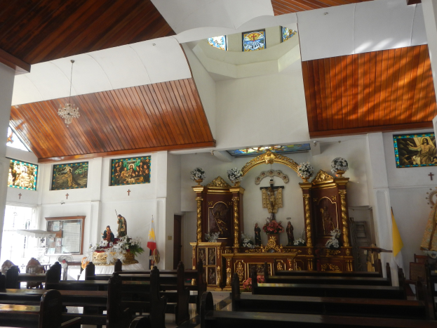 Innenseite einer Kirche mit Holzlehnen, Statuen, Blumenbouquets, Fahnen, gerahmten Fotos an den Wänden und Buntglasfenstern, beleuchtet von Deckenlampen.
