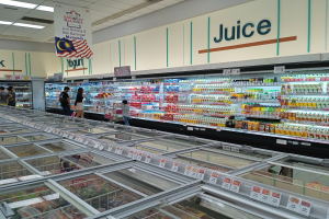 Ein hell erleuchteter Saftladen in einem Supermarkt zeigt eine Reihe von Flaschengetränken, frischem Gemüse und anderen Lebensmitteln auf Regalen, mit ein paar Menschen, die durch die Gänge schlendern und einer deckenmontierten Leuchttafel, die den Raum beleuchtet.