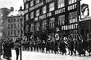 Ein Schwarz-Weiß-Bild einer Gruppe von Menschen, die eine Straße entlang marschieren und Fahnen und Schilder halten, mit einem Laternenpfahl links und einem Gebäude mit einer Tafel rechts, auf der 'Deutsche Nazi-Partei in Berlin' steht.