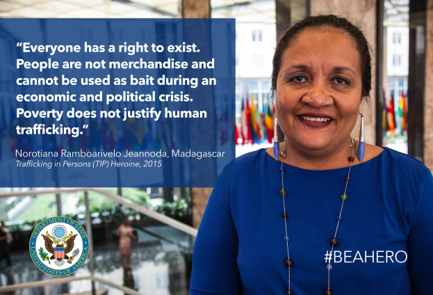 Eine Frau in einem blauen Kleid steht lachend vor einem Gebäude mit einem Zitat über Menschenhandel, umgeben von Menschen, Pflanzen und Flaggen.