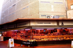 Ein belebter Markt mit verschiedenen Lebensmitteln auf einem Stand, beleuchtet von Deckenlampen, umgeben von Texttafeln, mit Gebäuden und einem klaren blauen Himmel im Hintergrund.