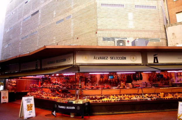 Ein belebter Markt mit verschiedenen Lebensmitteln auf einem Stand, beleuchtet von Deckenlampen, umgeben von Texttafeln, mit Gebäuden und einem klaren blauen Himmel im Hintergrund.