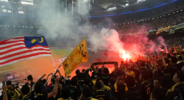 Eine gro├če Menschenmenge in einem Stadion mit Bengalos, Fahnen und einer Kamera, mit Rauch und Schildern, die "Malaysia vs Malaysia Live Streaming" im Hintergrund zeigen.