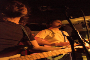 Eine Person in einem weißen Hemd singt in ein Mikrofon und spielt eine grüne Gitarre, während sie neben einer anderen Person in einem schwarzen Hemd und Brille steht, die ebenfalls singt und eine schwarze Gitarre spielt, mit Scheinwerfern und anderen Instrumenten im Hintergrund.