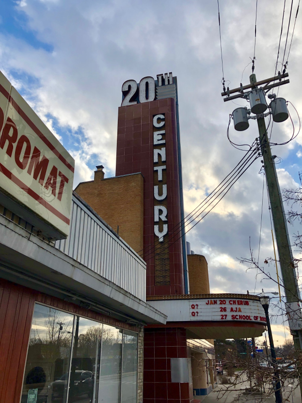 Außenansicht des 20th Century Theatre in Detroit, Michigan, mit Glaswänden und Namensschildern, umgeben von Bäumen, Strommasten und Drähten im Vordergrund und einem bewölkten Himmel im Hintergrund.