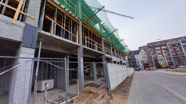 Großer Bau auf einer Stadtstraße mit Fahrzeugen, Straßenlaternen und umliegenden Gebäuden unter einem klaren blauen Himmel.