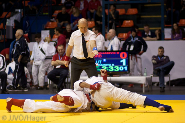 Zwei Judoka im Kampf mit einem Schiedsrichter in der Nähe, Zuschauern im Hintergrund und einem Bildschirm mit Match-Informationen.
