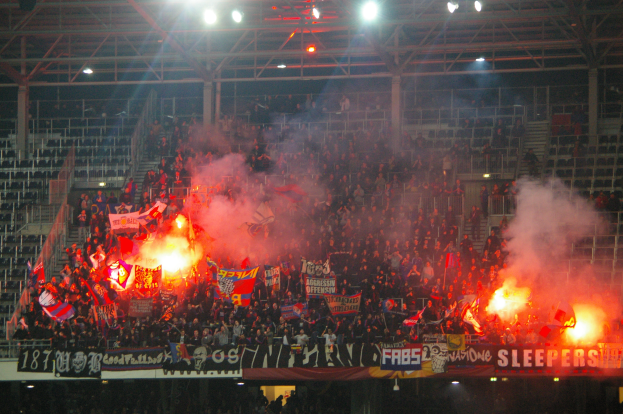 Eine große Menge Menschen in einem Stadion hält Fahnen und Banner, mit Leuchtraketen, die Rauch erzeugen, unter einem Dach mit Deckenleuchten und Metallrahmen.