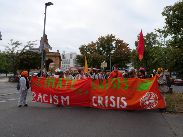 Gruppe von Menschen marschiert auf einer von Bäumen gesäumten Straße mit einem "Klimakrise ist eine Krise"-Schild, Fahrzeuge fahren vorbei, Gebäude und klarer blauer Himmel im Hintergrund.