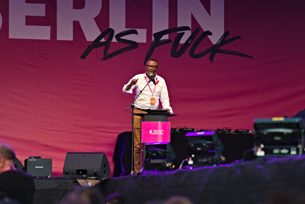 Ein Mann in weißem Hemd und braunen Hosen steht auf einer Bühne und hält ein Mikrofon in der Hand, mit einem pinken Banner mit der Aufschrift 'Berlin as Fuck' im Hintergrund und Lautsprechern auf der Bühne.
