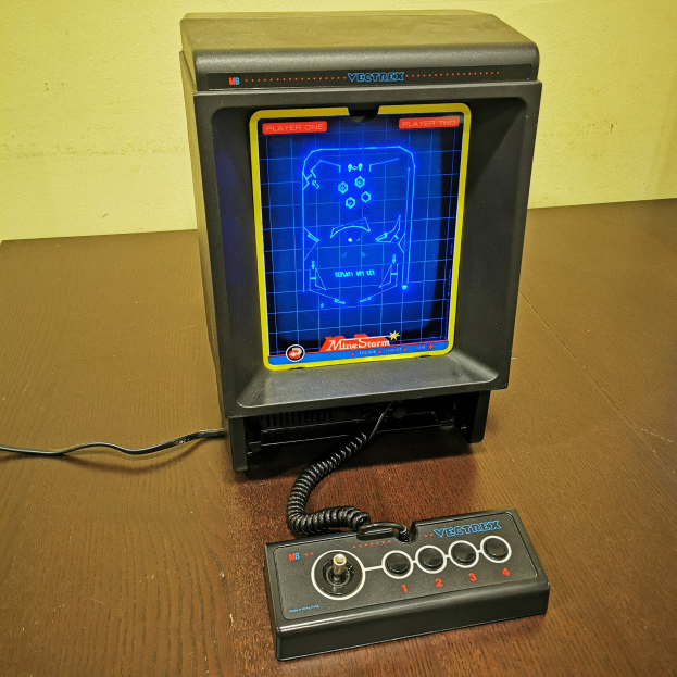 Alte Video-Spielkonsole auf einem Holztisch mit einem angeschlossenen Kabel und einer Wand im Hintergrund.