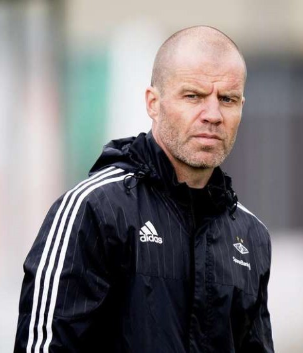 Mann mit Glatze in schwarzer Jacke, marineblauer Jacke mit weißen Streifen und weißem Kragen, der auf einem Fußballfeld steht und die Hände zusammengelegt hat.