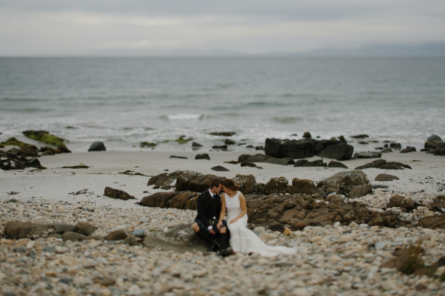 Bräutigam und Braut umarmend und küssend auf einem felsigen Strand mit Meer und Himmel im Hintergrund.