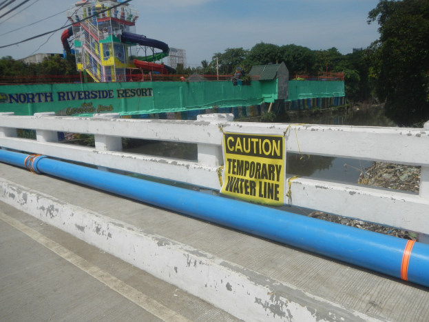 Vorsichtsschild mit der Aufschrift "Vorsicht temporäre Wasserleitung" auf einer Brücke mit Gelönder, Bäumen, Gebäuden, einer Wasserrutsche und einem bewölkten Himmel im Hintergrund.